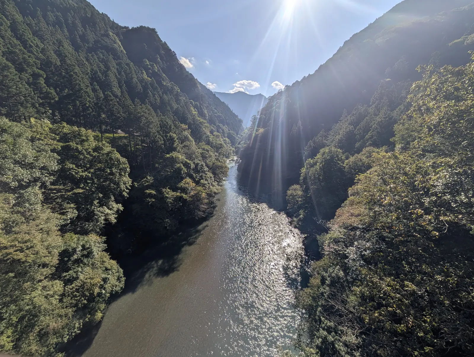 逆光で撮った川の谷。画面上部の太陽から光芒が放射状に伸び、川面は一面のまぶしい反射になり、両側の斜面は暗く沈んでいる。